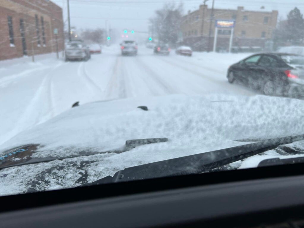 View from inside a vehicle driving down a snowy city street during a winter storm, with snow covering the hood and windshield.