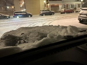 View from inside a vehicle at night, looking over a snow-covered hood onto a snowy city street lined with parked cars.