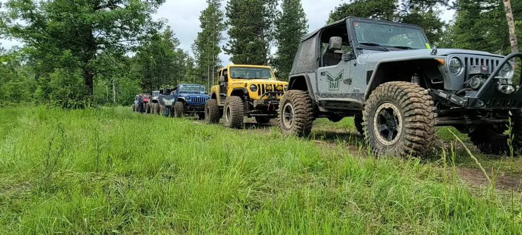 A line of off-road vehicles, predominantly Jeep models, is parked on a lush green trail surrounded by trees. The vehicles are equipped with large, rugged tires suitable for off-roading. The yellow Jeep in the middle of the line and a gray Jeep with TNT Customs decals in the foreground stand out prominently. These Jeeps are lined up for a TNT Customs Event.