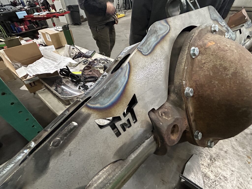 Close-up of a TNT Customs axle truss installed on a Jeep axle, featuring precision welds, a laser-cut TNT logo, and durable construction in a workshop setting.