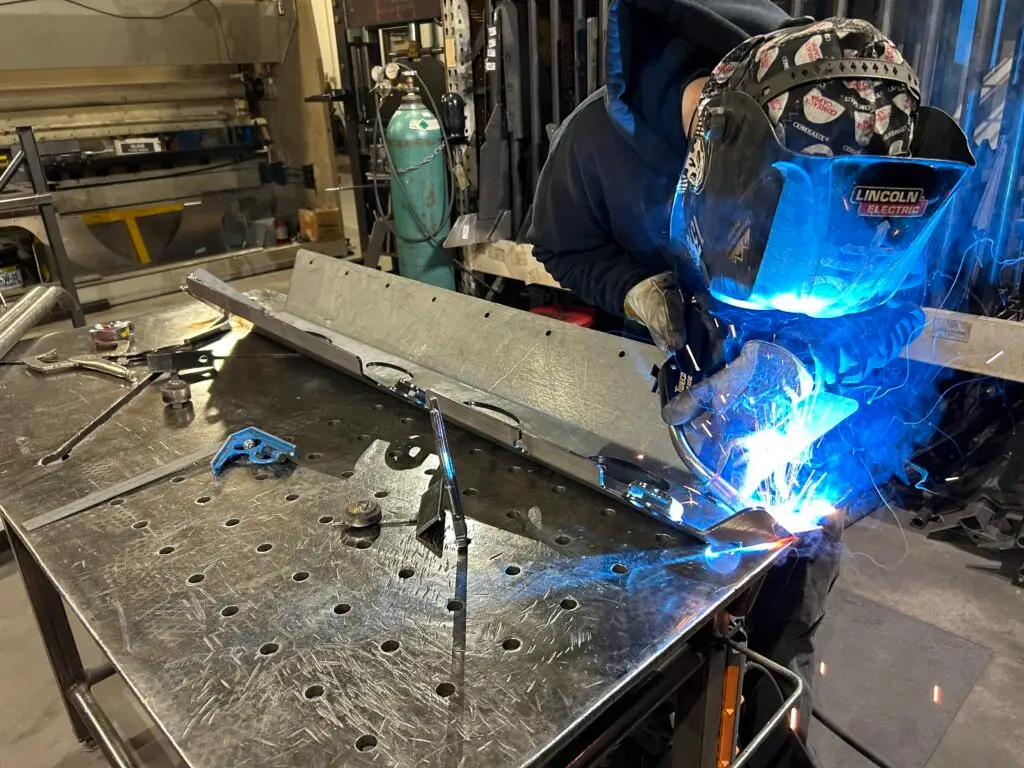 A TNT Customs fabricator welding a Jeep aftermarket part in the workshop, with sparks flying as they work on a metal component. The fabricator is wearing a protective welding helmet and gloves.
