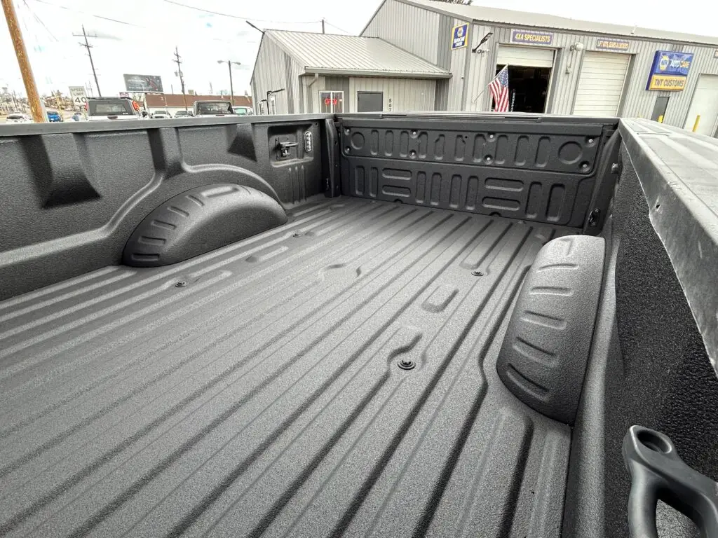 Close-up view of a truck bed featuring a Mil-Spec spray-on bedliner, applied with a textured, durable coating for added protection. The image is taken outdoors near TNT Customs, with their shop and signage visible in the background.
