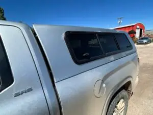 A silver Toyota Tacoma with an ARE truck topper installed, parked in a dirt lot near a red industrial building.