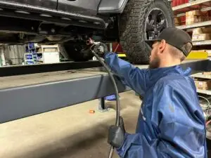 A TNT Customs technician in protective gear sprays undercoating onto the underside of a lifted Jeep inside a workshop, ensuring rust and corrosion protection.