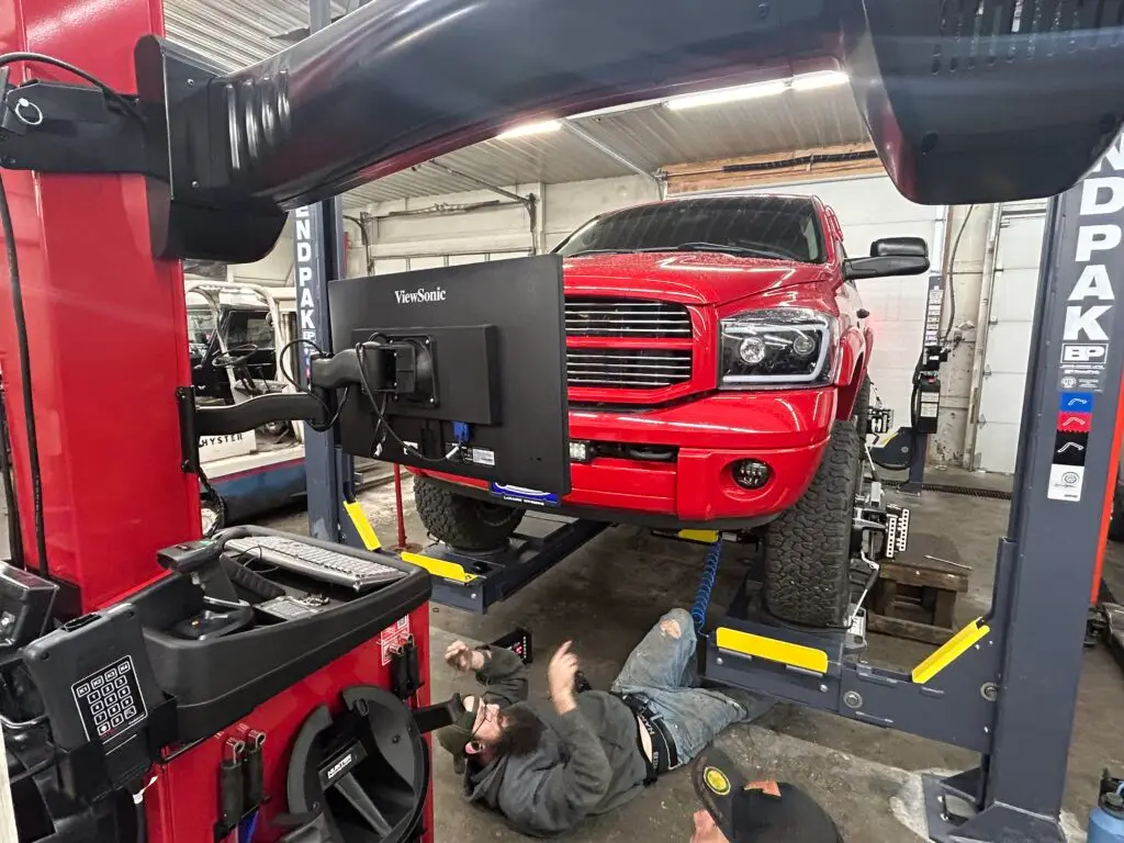 A technician working under a red truck on an alignment lift, with a computer screen showing alignment diagnostics.