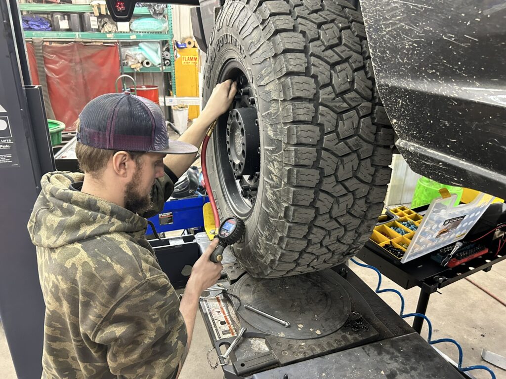 Technician inflating a large all-terrain tire with a digital pressure gauge inside the TNT Customs shop.