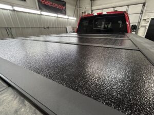 Close-up of a black textured tri-fold tonneau cover installed on a red pickup truck inside an auto shop.