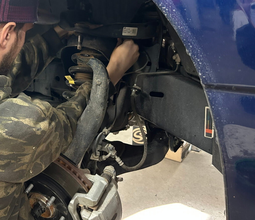 Technician installing suspension components on a lifted truck at TNT Customs, working inside the wheel well on a vehicle lift upgrade.
