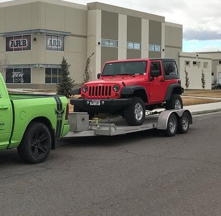 Green pickup truck towing a red Jeep Wrangler on a dual-axle car hauler trailer.