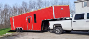 White pickup truck towing a large red gooseneck enclosed trailer with triple axles parked on a driveway.